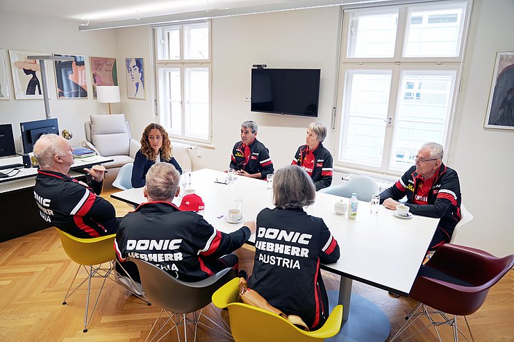 Aufnahme eines Tisches, an dem sieben Männer und Frauen sitzen. 