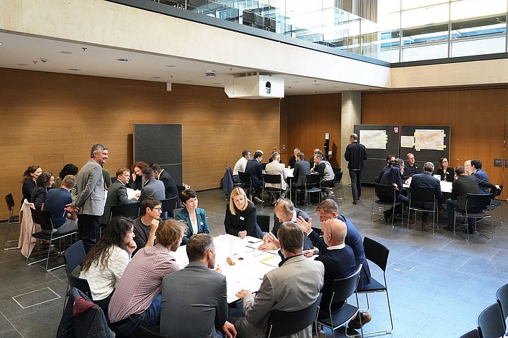 Menschen in Saal um große Tische sitzend und diskutierend