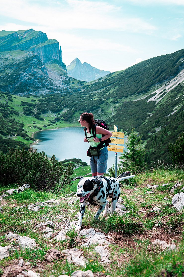 © Marco Christanell Eine Frau mit Hund auf einem Wanderweg im Hintergrund ein Bergsee und gelbe Hinweisschilder