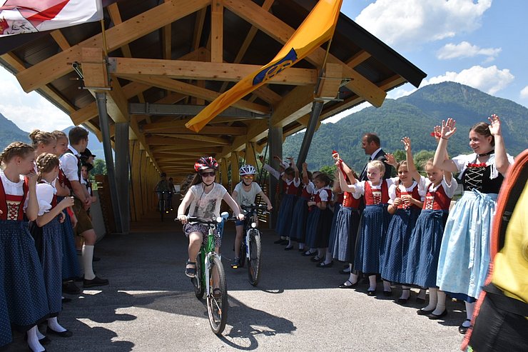Zwei SchülerInnen fahren mit dem Rad über die Brücke und werden von Kindern des Trachtenvereins Erl beklatscht.