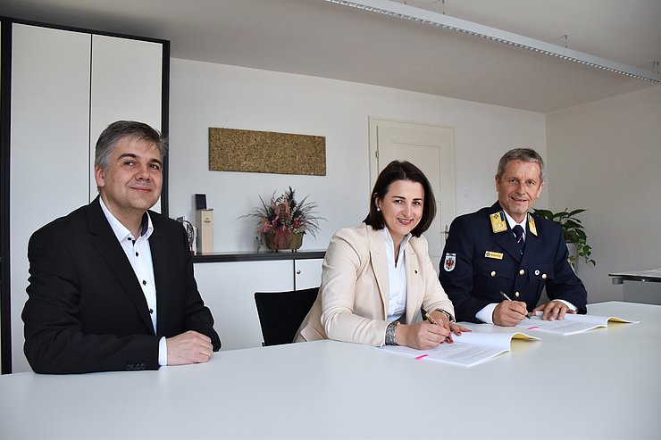 LRin Mair und Stock am Tisch sitzend Vertrag unterschreibend, daneben Reister sitzend