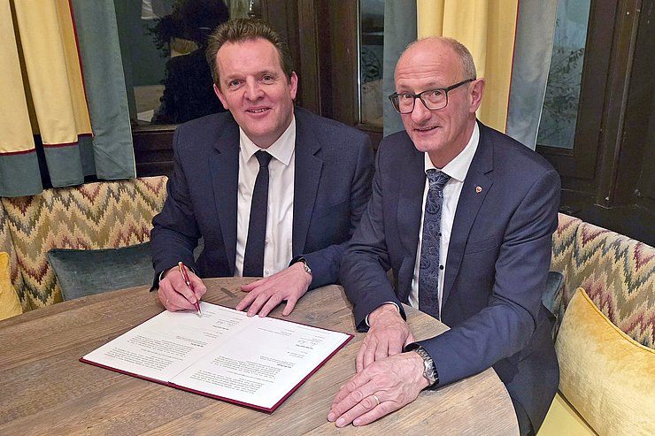 © Land Tirol Zwei Männer sitzen an einem Tisch, der linke unterzeichnet einen Vertrag.