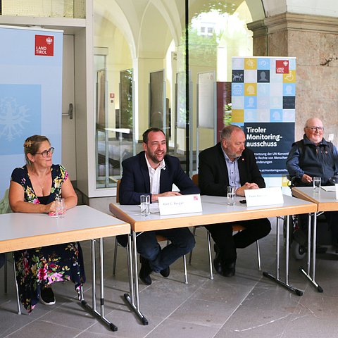 Tisch mit RednerInnen, im Hintergrund 2 Roll-ups des Landes/Monitoringausschusses