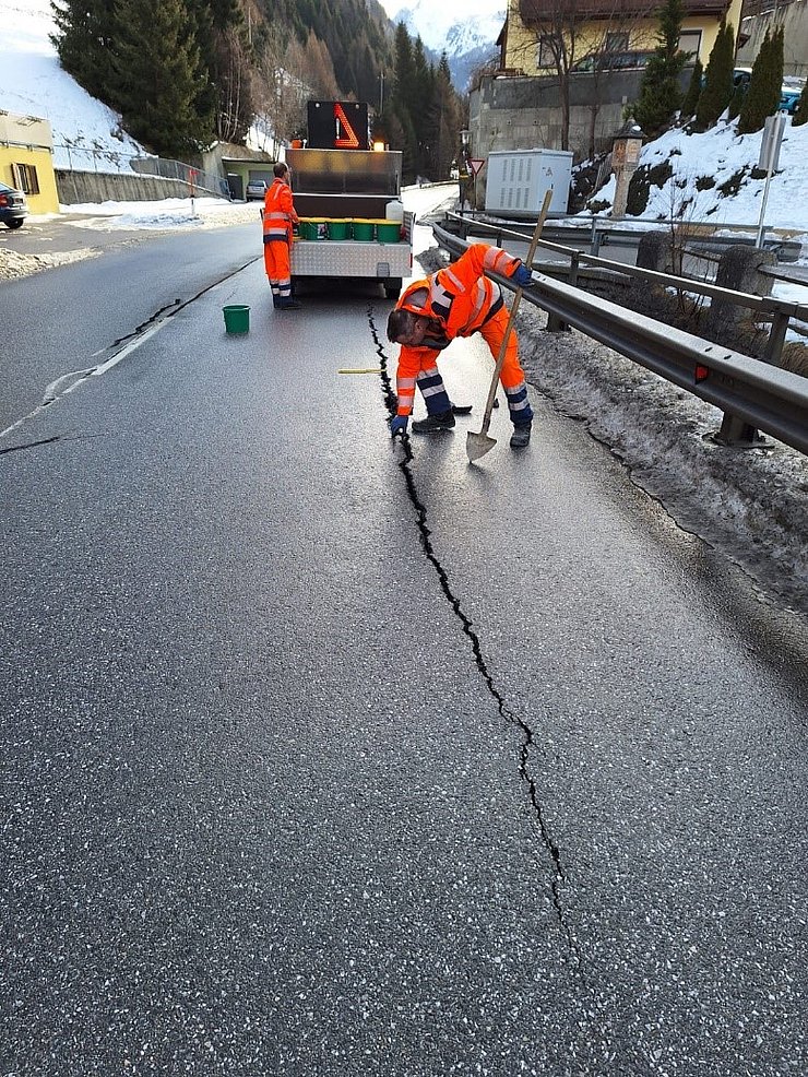 © Land Tirol Straße mit Riss, Mann in orangen Overall mit Schaufel beugt sich über Riss