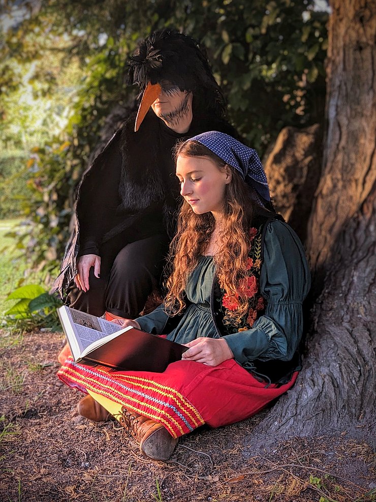 Ein Mädchen mit Kopftuch sitzt unter einem Baum und liest, daneben kniet ein als Rabe verkleideter Mann. 