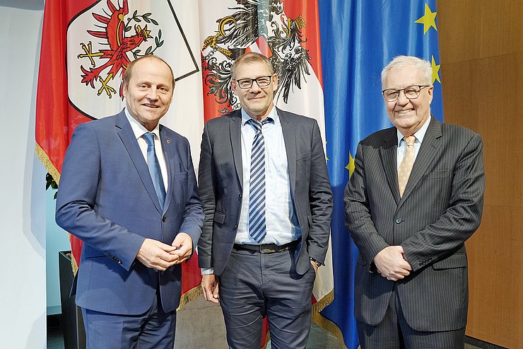Drei Männer in Anzügen stehen vor Fahnen in einem Saal. 