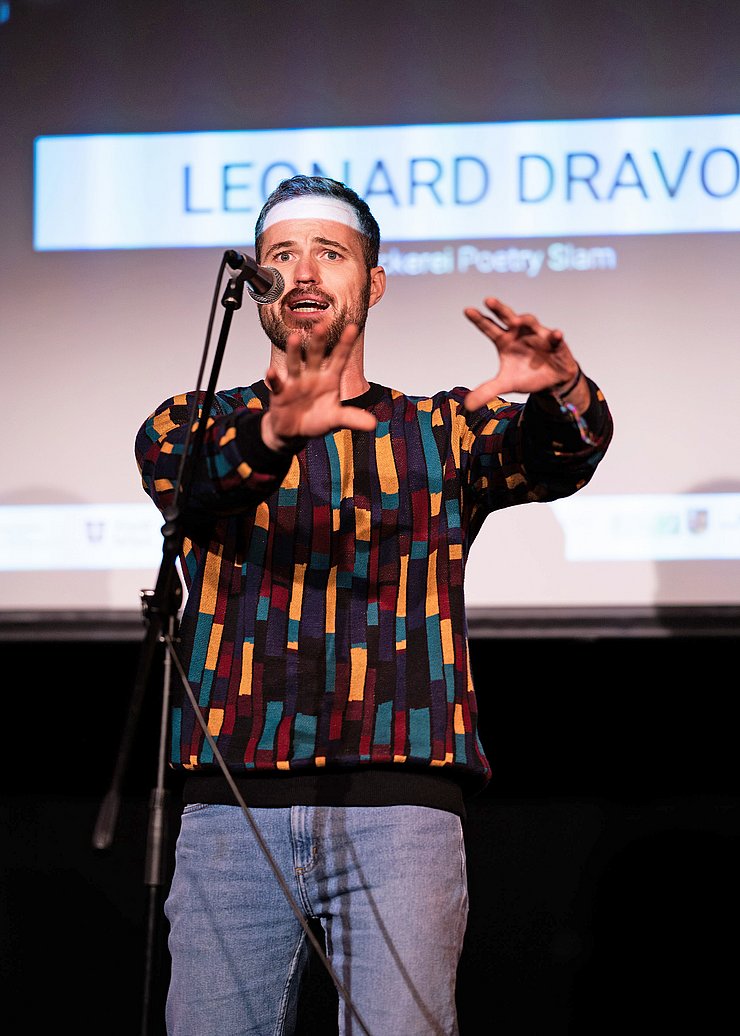 © Yasmin Abbas Ein Mann steht auf einer Bühne, spricht in ein Mikrofon und gestikuliert mit beiden Händen.