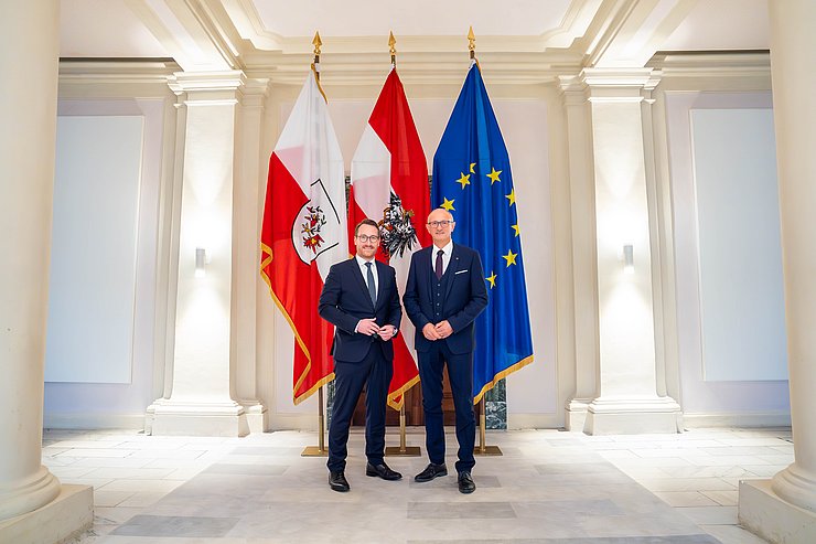 Zwei Männer stehen vor Fahnen und blicken in die Kamera.