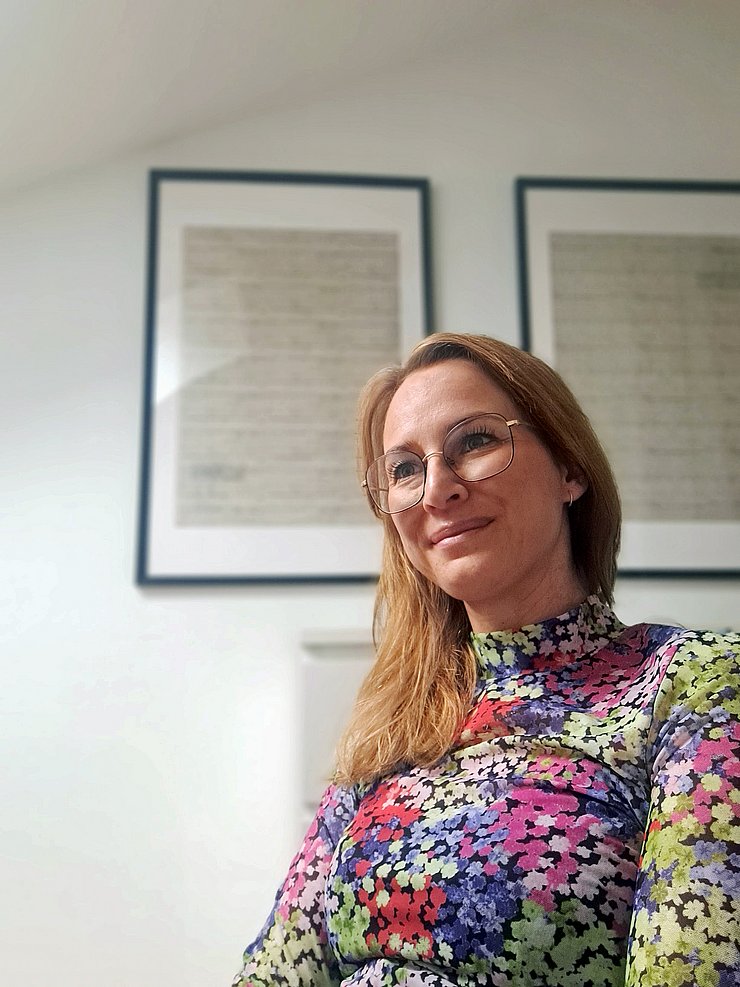 © Privat/Magdalena Schweissgut Eine Frau mit Brille sitzt vor einer Wand mit zwei Bildern.