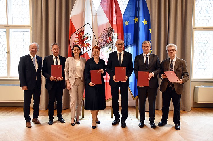 Sieben Personen stehen vor drei verschiedenen Fahnen und halten unterzeichnete Vereinbarungen in ihren Händen.