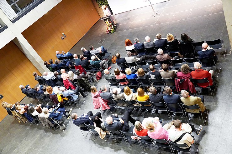 Luftaufnahme des Großen Saals im Landhaus, viele Personen sitzen auf Stühlen nebeneinander und hören einer Rednerin zu.