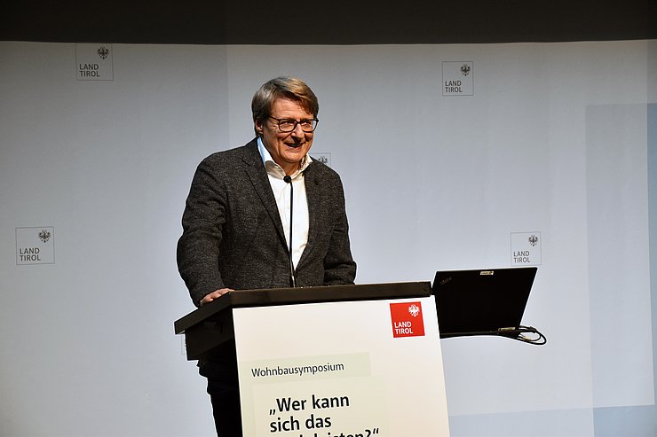 Gschwentner hinter Rednerpult stehend; Rednerpult mit Sujet von Symposium "Wer kann sich das noch leisten?"