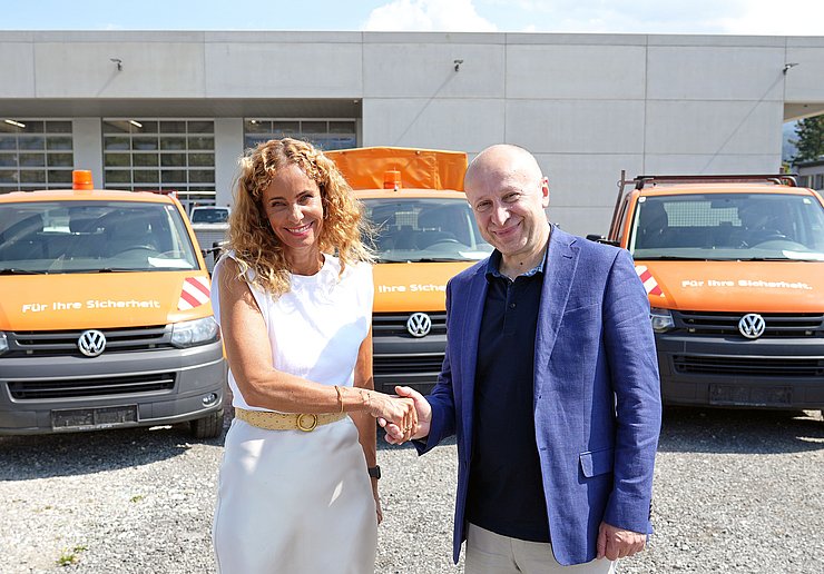 Eine Frau schüttelt einem Mann die Hand, im Hintergrund sind mehrere Fahrzeuge zu sehen.