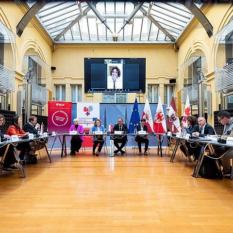 Personen sitzen an U-förmigem Tisch, darüber Bildschirm