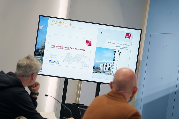 © Land Tirol/Neuner Blick zwischen zwei Männer hindurch auf eine Tafel.