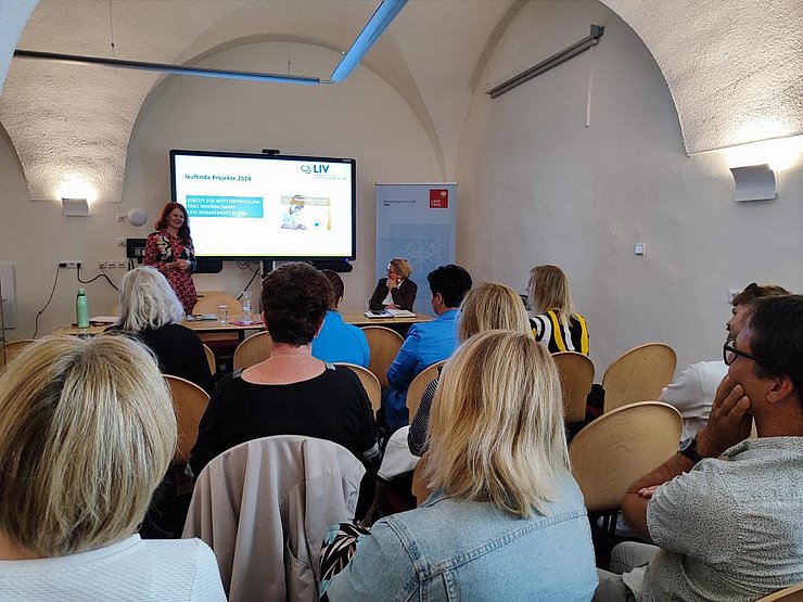 Foto von Personen in einem Raum mit runden Bogen an der Decke. Personen sind von hinten zu sehen während sie zwei weiteren Personen die vor ihnen stehen und sitzen zuhören.