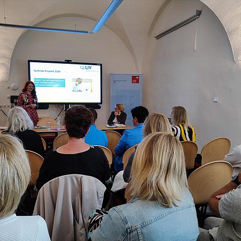 Foto von Personen in einem Raum mit runden Bogen an der Decke. Personen sind von hinten zu sehen während sie zwei weiteren Personen die vor ihnen stehen und sitzen zuhören.