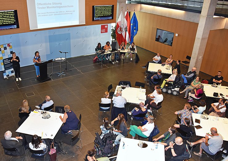 Foto auf den Saal von oben: es stehen verteilt weiße Tische im Raum, dort sitzen die TeilnehmerInnen; vorne spricht Kafka, neben ihr die Gebärdendolmetscherin