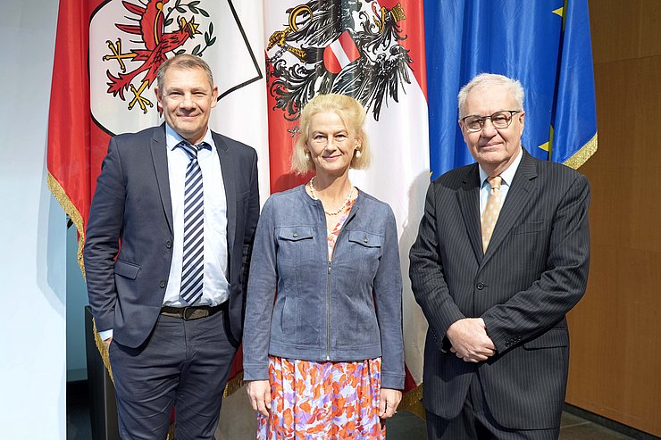 Zwei Männer im Anzug stehen vor Fahnen, zwischen ihnen steht eine blonde Frau. 