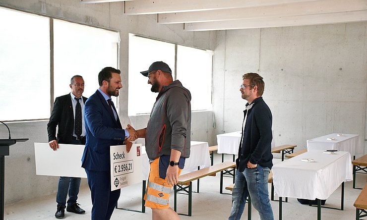 © Land Tirol/Hörmann Dornauer mit großem Scheck in der Hand schüttelt Bauarbeiter die Hand