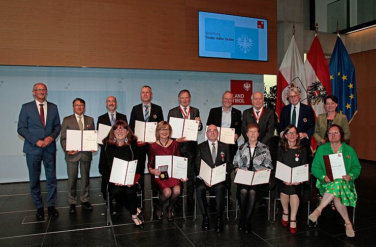 13 Persönlichkeiten, die mit dem Tiroler Adler-Orden geehrt wurden, sitzen oder stehen in einer Gruppe im Landhaus, gemeinsam mit LH Mattle (li.) und LTPin Ledl-Rossmann (re.).