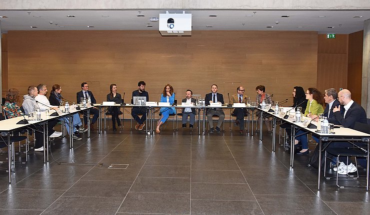 Foto vom Saal mit U-förmigem Tisch, an dem Personen sitzen