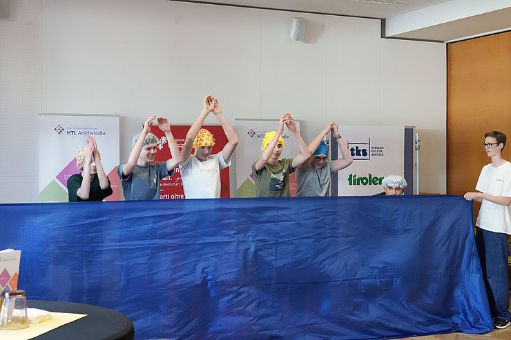 SchülerInnen mit Bademasken heben Hände hoch, davor blaues Tuch (steht für Wasser)