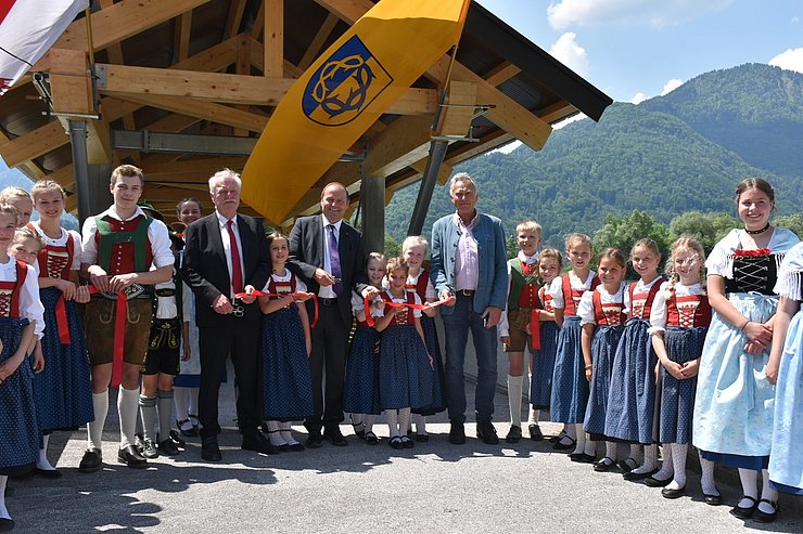 Ein Foto von Bgm von Erl Georg Aicher-Hechenberger, LHstv. Josef Geisler und 2. Bgm von Oberaudorf Alois Holzmaier gemeinsam mit den Kindern des Trachtenvereins Erl bei der Eröffnungsfeier des neugebauten Innstegs Erl vor der Brücke beim symbolischen durchschneiden des roten Bandes
