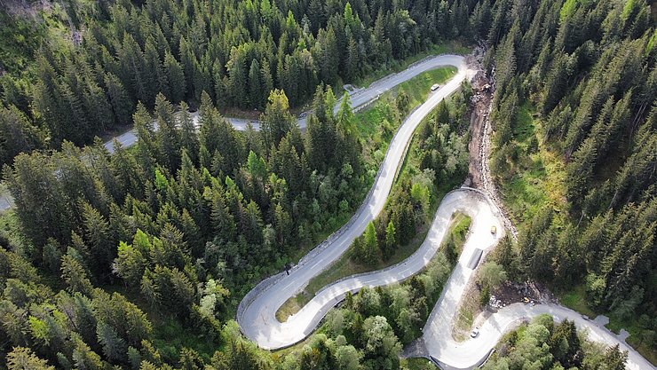 Lutaufnahme der Straße mit vielen Kehren durch den Wald; an einer Kehre sieht man das Bachbett, dass teilweise auf die Straße getreten ist