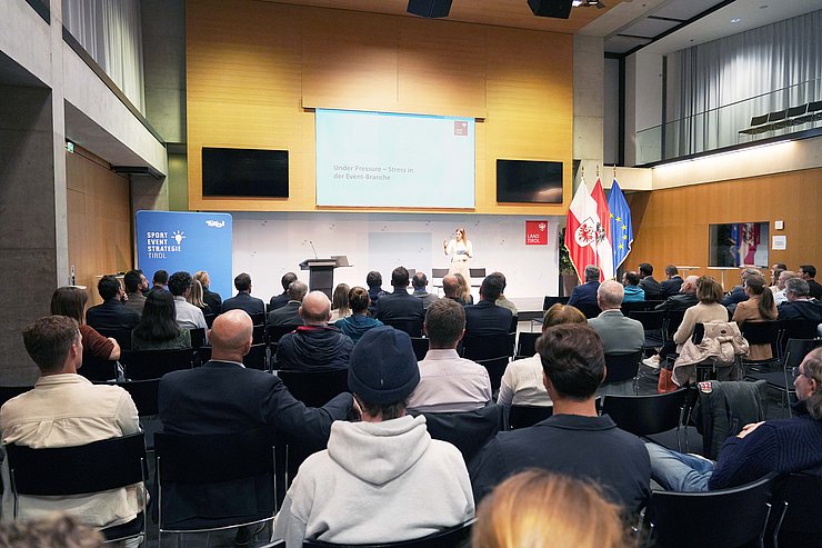 Großer Saal im Landhaus: Menschen sitzen in Stuhlreihen; auf Bühne spricht Frau