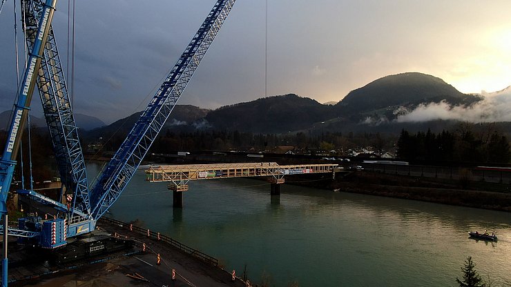 Ein Foto in der Dämmerung, welches sowohl den 800 Tonnen schweren Kran als auch die sich in Umbau befindliche Rad- und Fußgängerbrücke über den Inn zeigt. 
