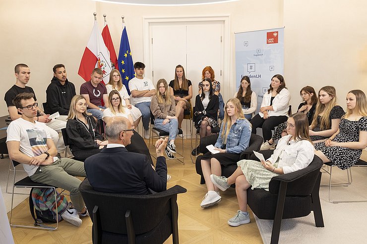© Land Tirol/Die Fotografen Die Schülerinnen und Schüler sitzen auf Stühlen im Halbkreis um Landeshauptmann Mattle