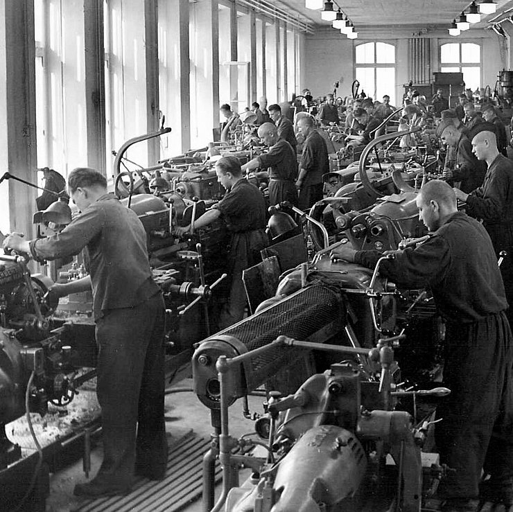 © Gemeindearchiv Kematen In einheitlicher Arbeitskleider stehen jeweils Männer vor einer Drehbank in einer Fabrikshalle.