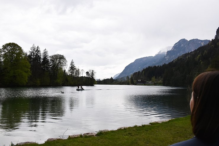 © Land Tirol/Pölzl Rechts am Bild der Hinterkopf von Mair; sie blickt auf den Reintalersee vor ihr. Im See schwimmt eine Person und winkt um Hilfe. Ein Boot mit Mitgliedern der Wasserrettung nähert sich. Im Hintergrund der wolkenverhangene Himmel und Berge