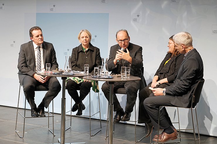 Mehrere Personen sitzen rund um einen Tisch und führen ein Gespräch mit einem Mikro. 