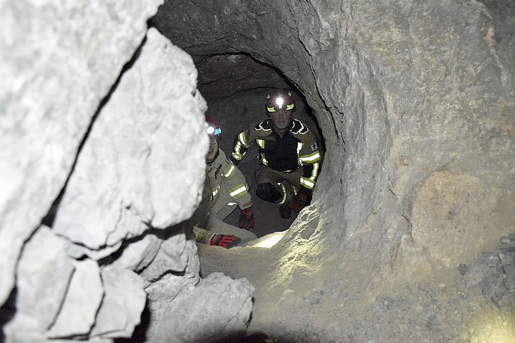 ein enges rundes LOck (ca. einen Meter groß); unten stehen zwei Mitglieder der Rgubenwehr und blicken hoch, Auf ihren Helmen sind Stirnlampen, die in die Kamera leuchten