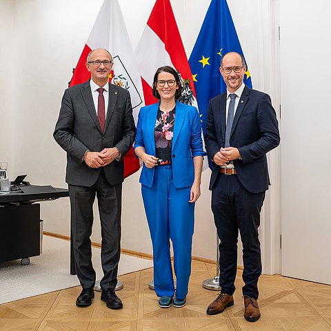 Barbara Thaler im blauen Hosenanzug steht im Büro Landeshauptmann vor den Fahnen Tirols, Österreichs und der EU: An ihrer linken Seite LH Mattle, an ihrer rechten Seite LR Gerber, beide im dunklen Anzug mit Krawatte.