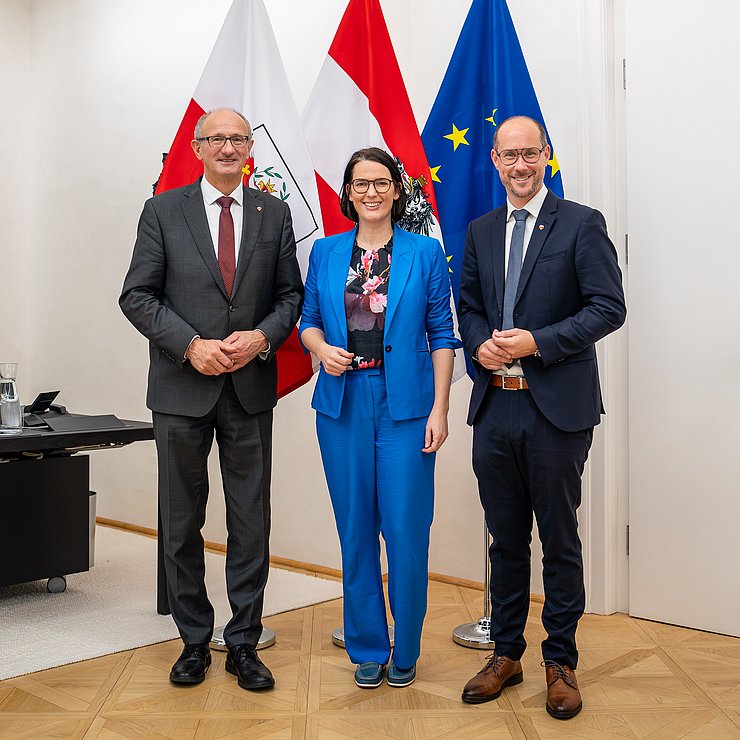 Barbara Thaler im blauen Hosenanzug steht im Büro Landeshauptmann vor den Fahnen Tirols, Österreichs und der EU: An ihrer linken Seite LH Mattle, an ihrer rechten Seite LR Gerber, beide im dunklen Anzug mit Krawatte.