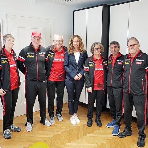 Sechs Männer und Frauen stehen im Sportanzug neben einer elegant gekleideten Frau. 