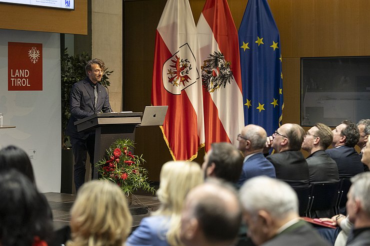 © Land Tirol/Die Fotografen Ein Mann steht an einem Rednerpult, vor ihm sitzen mehrere Leute im Saal.