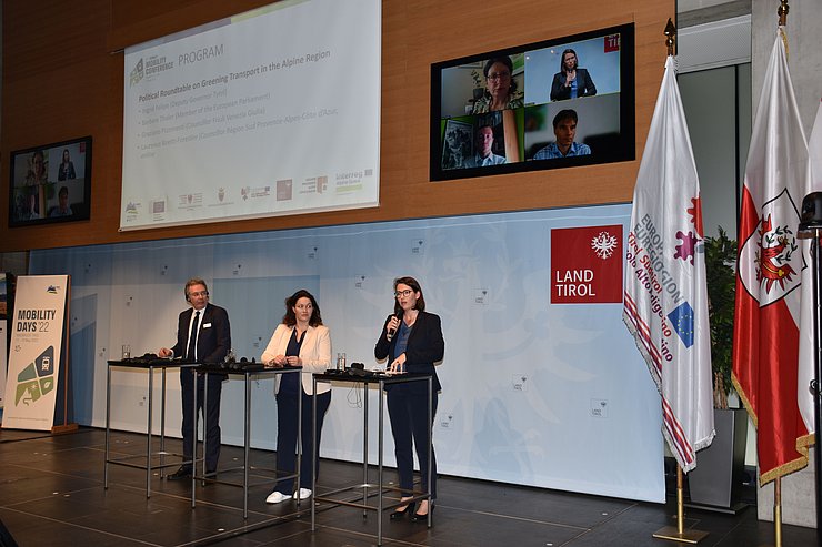 © Land Tirol/Dorfmann Die GesprächsteilnehmerInnen an den Sprechpulten, darüber ein Bildschirm mit den online zugeschalteten TeilnehmerInnen.