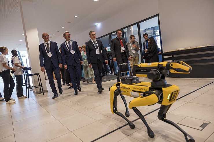 © Land Tirol/Sedlak Ein Roboter im Vordergrund, im Hintergrund gehen Männer im Anzug.