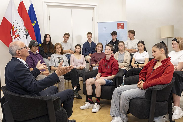 © Land Tirol/Die Fotografen Die Klasse sitzt im Halbkreis vor LH Anton Mattle und hört ihm zu.