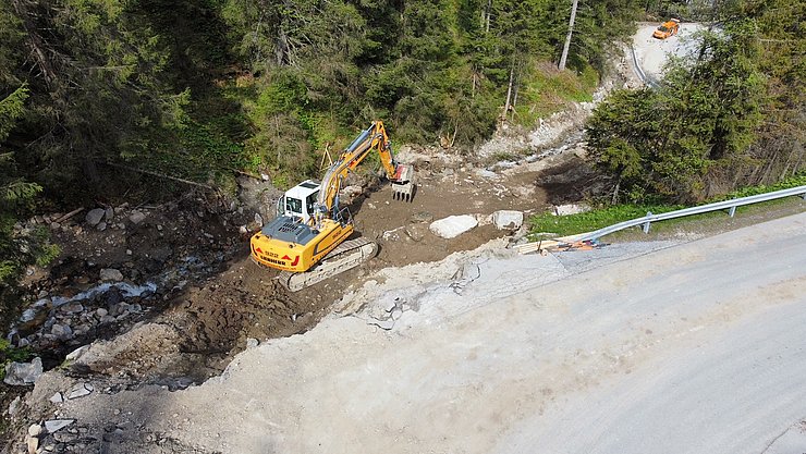Das Bachbett neben der Straße ist teilweise auf die Straße getreten; im leeren Bachbett steht ein gelber Bagger