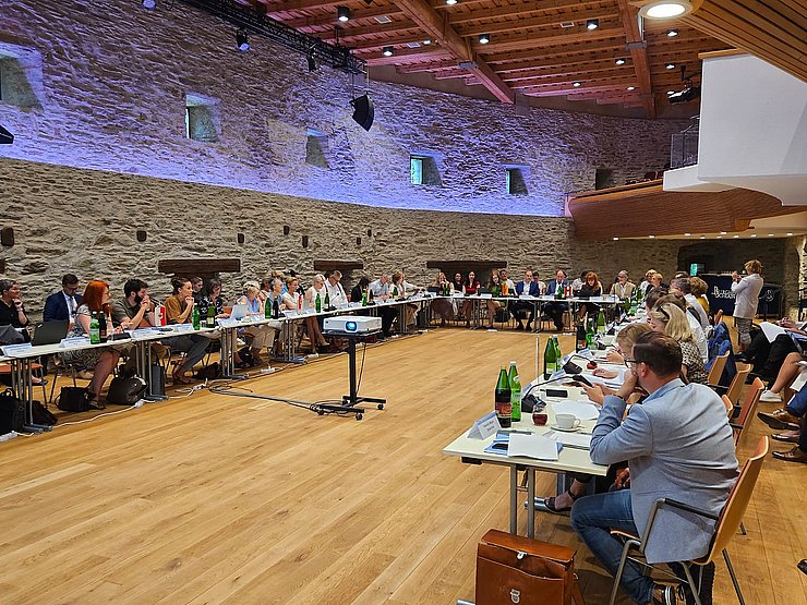 Saal mit Tisch in U-Form, an dem Personen sitzen, dahinter blau beleuchtete Steinwand