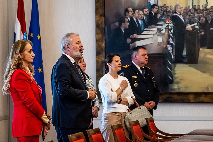 © Land Tirol/Christanell Mair, Sanader, eine Frau im roten Hosenanzug und ein kroatischer Feuerwehrmann im Parlament. Im Hintergrund ein riesen Gemälde mit einer Szene aus dem Parlament