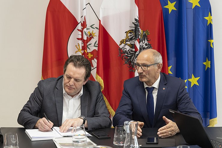 © Land Tirol/Die Fotografen LR Zumtobel und LH Mattle sitzen nebeneinander auf einem Tisch und diskutieren mit anderen Anwesenden.