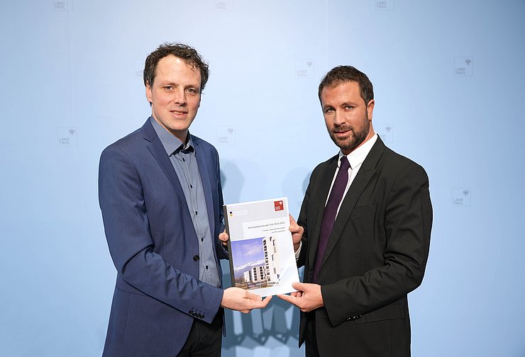 © Land Tirol/Neuner Zwei Männer blicken in die Kamera und halten eine Mappe.