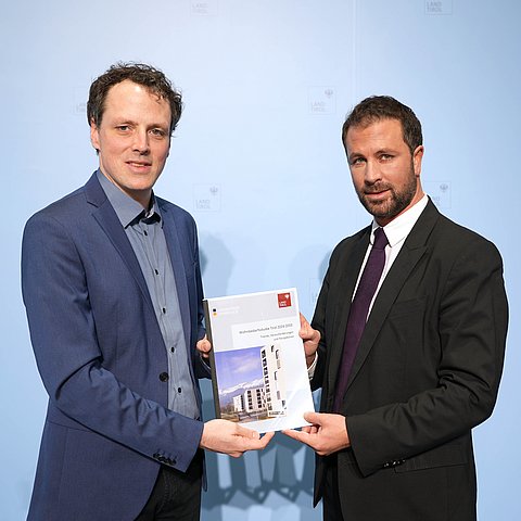 Zwei Männer blicken in die Kamera und halten eine Mappe.