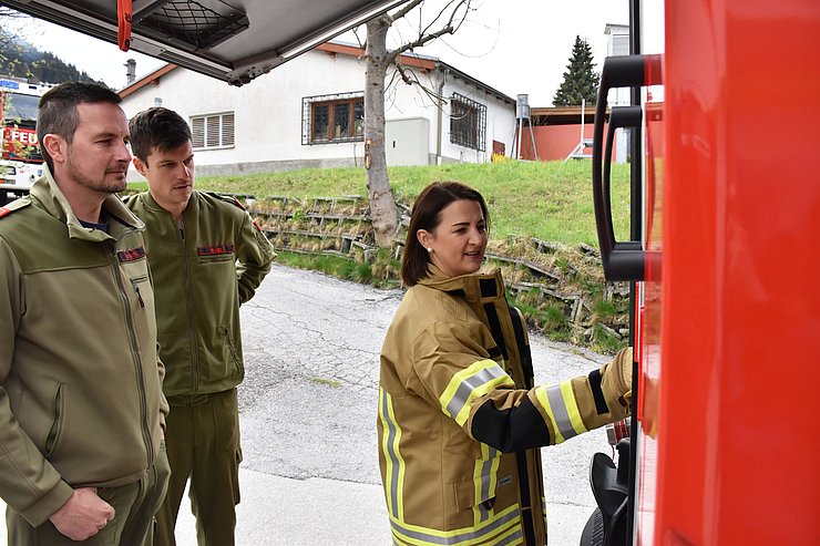 Mair mit gelber Feuerwehr-JAcke steht vor rotem Feuerwehr-Fahrzeug und greift auf Amatur. Links neben ihr stehen Muigg und sein Stellvertreter, beide in grünen Feuerwehruniformen und sehen Mair zu. Im Hintergrund ist ein Haus zu sehen und ein weiteres rotes Feuerwehr-Fahrzeug.